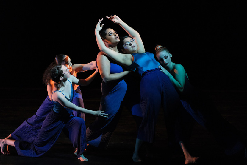 Five ballet performers in dresses leaning against each other on a dark background